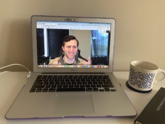 A man on a laptop screen during a video call. The laptop is on a desk with a mug of coffee next to it